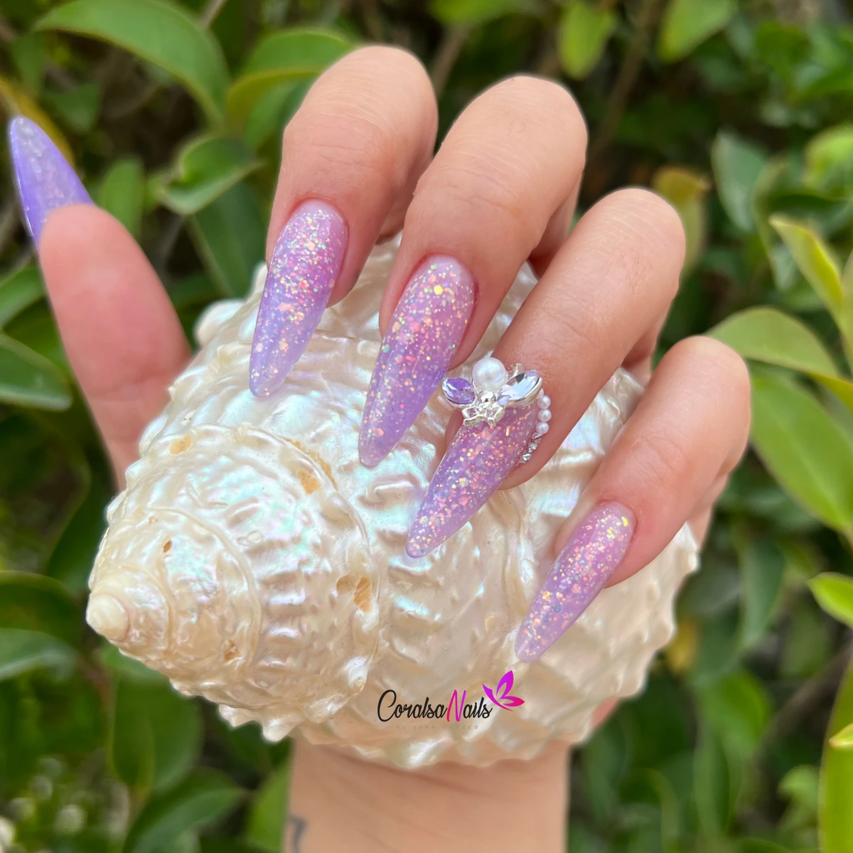Uñas moradas con brillo y decoración, sosteniendo una concha