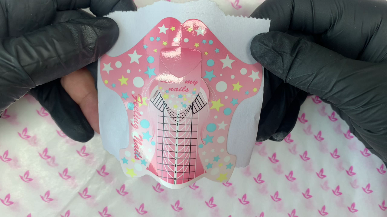 Molde adhesivo rosa para esculpir uñas, abierto y sostenido con guantes negros antes de adaptarlo.